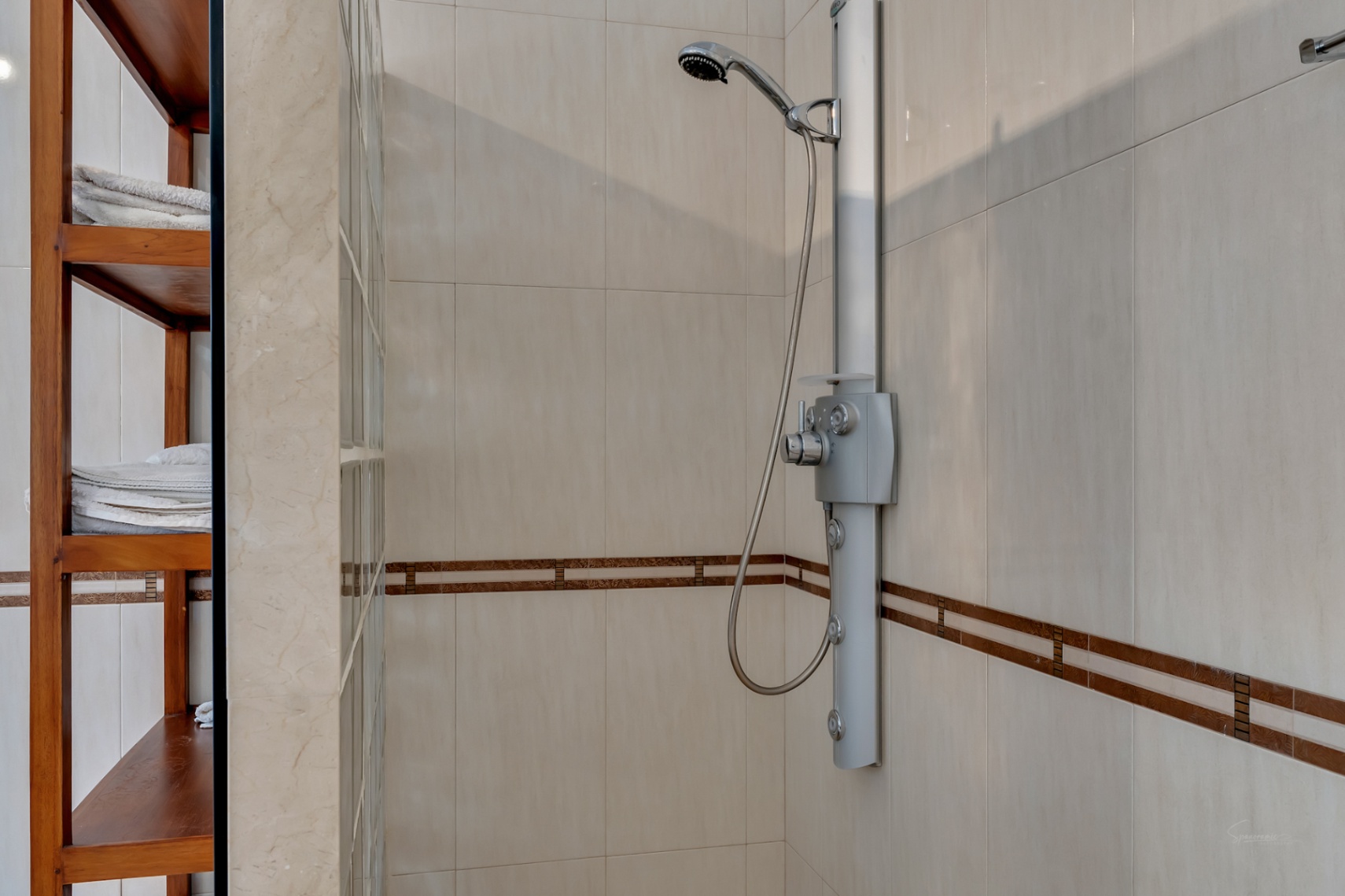 Douche moderne avec colonne d'hydromassage, carrelage crème avec frise décorative et étagères en bois intégrées près d'une paroi en briques de verre.