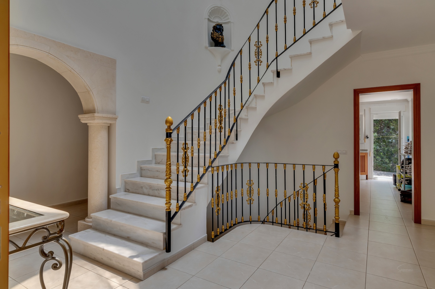 Escalier en marbre avec rampe en fer forgé et ornements dorés, jouxtant une arche en pierre de taille et une colonne dans le hall d'entrée.