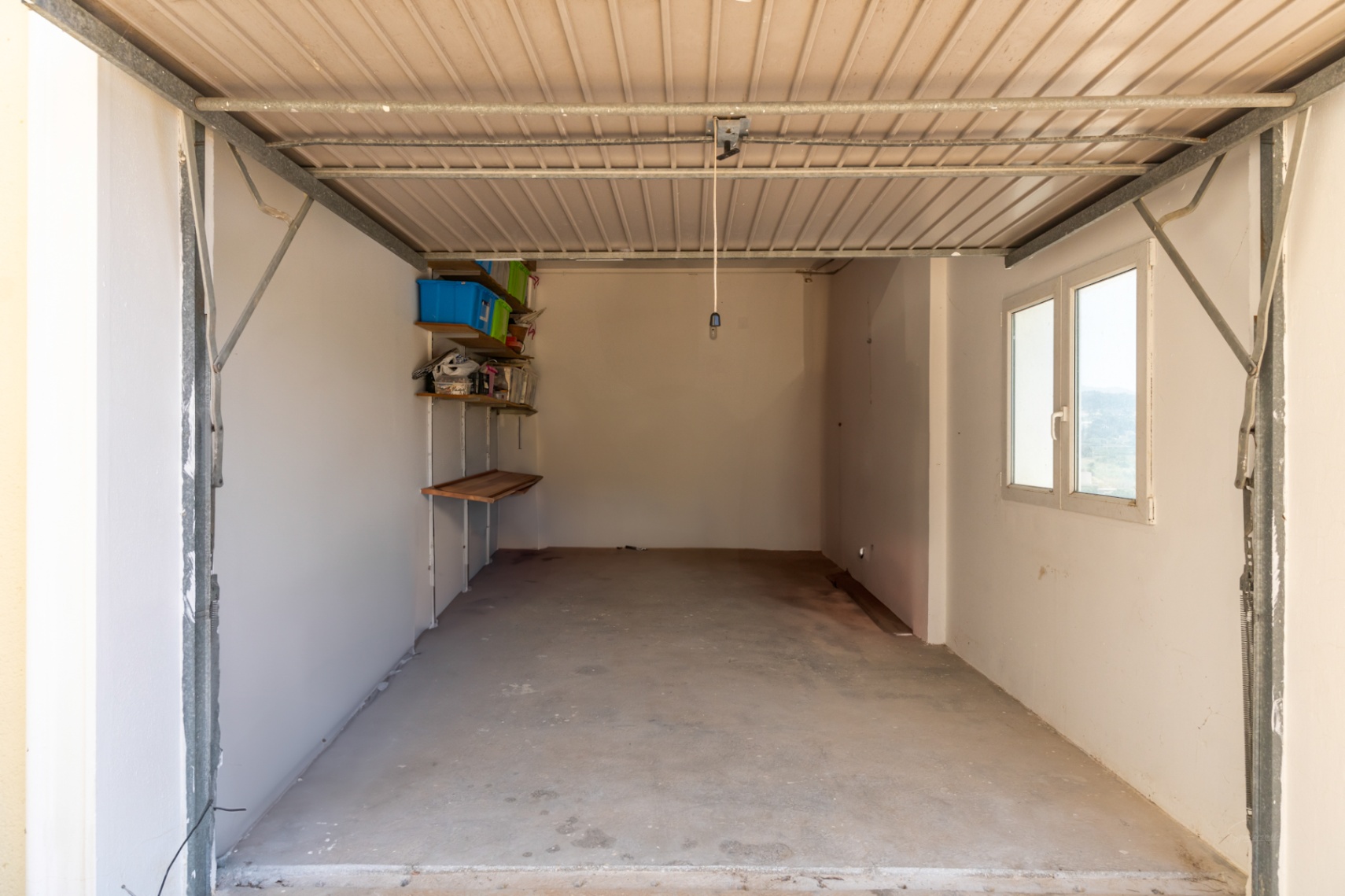 Garage privé avec sol en béton, murs blancs et fenêtre latérale. Comprend des étagères de rangement murales et une porte métallique basculante.