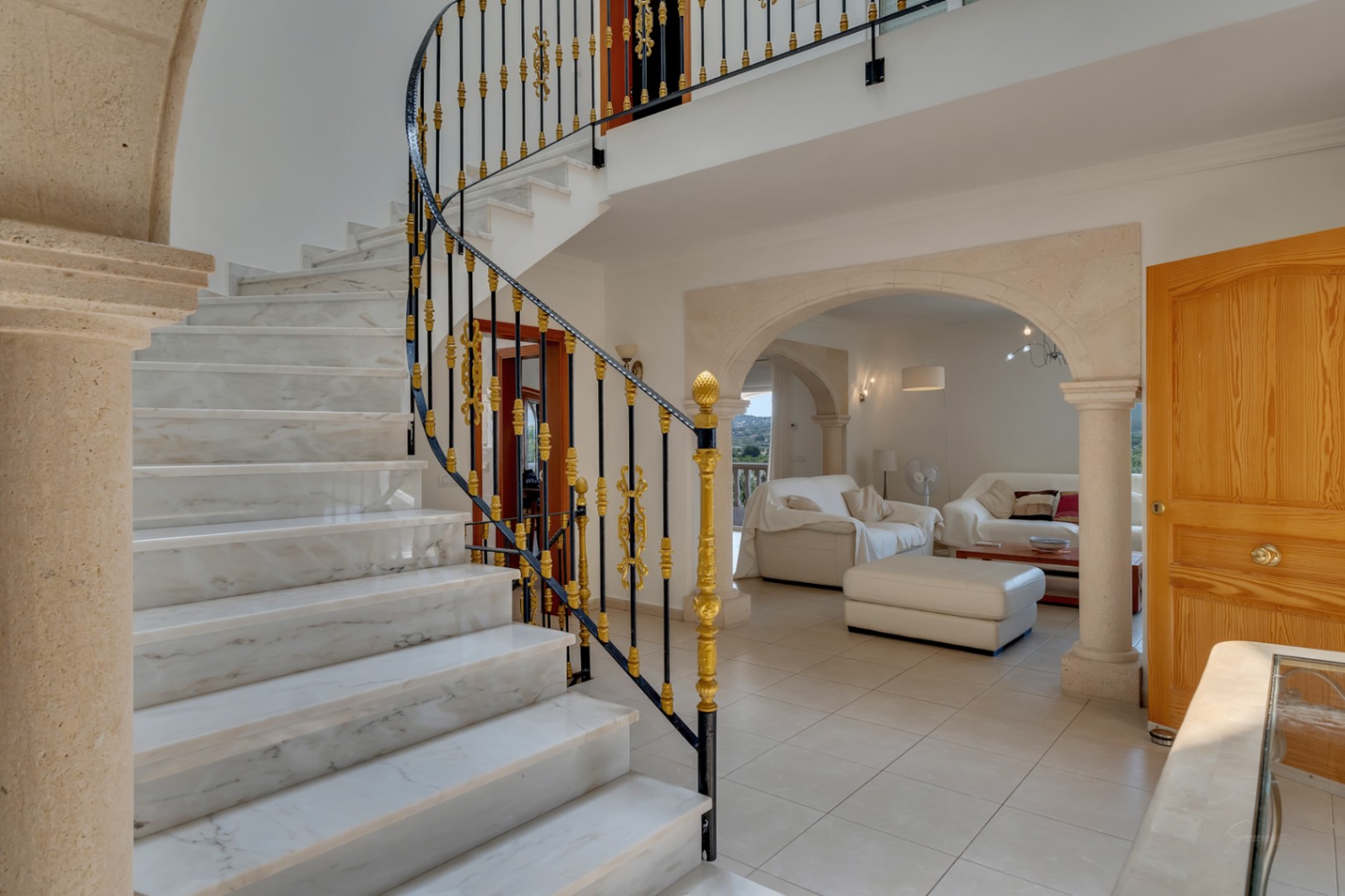 Hall d'entrée avec escalier en marbre et rampe ornementale, s'ouvrant sur un salon via des arches en pierre de Tosca et un sol en grès.