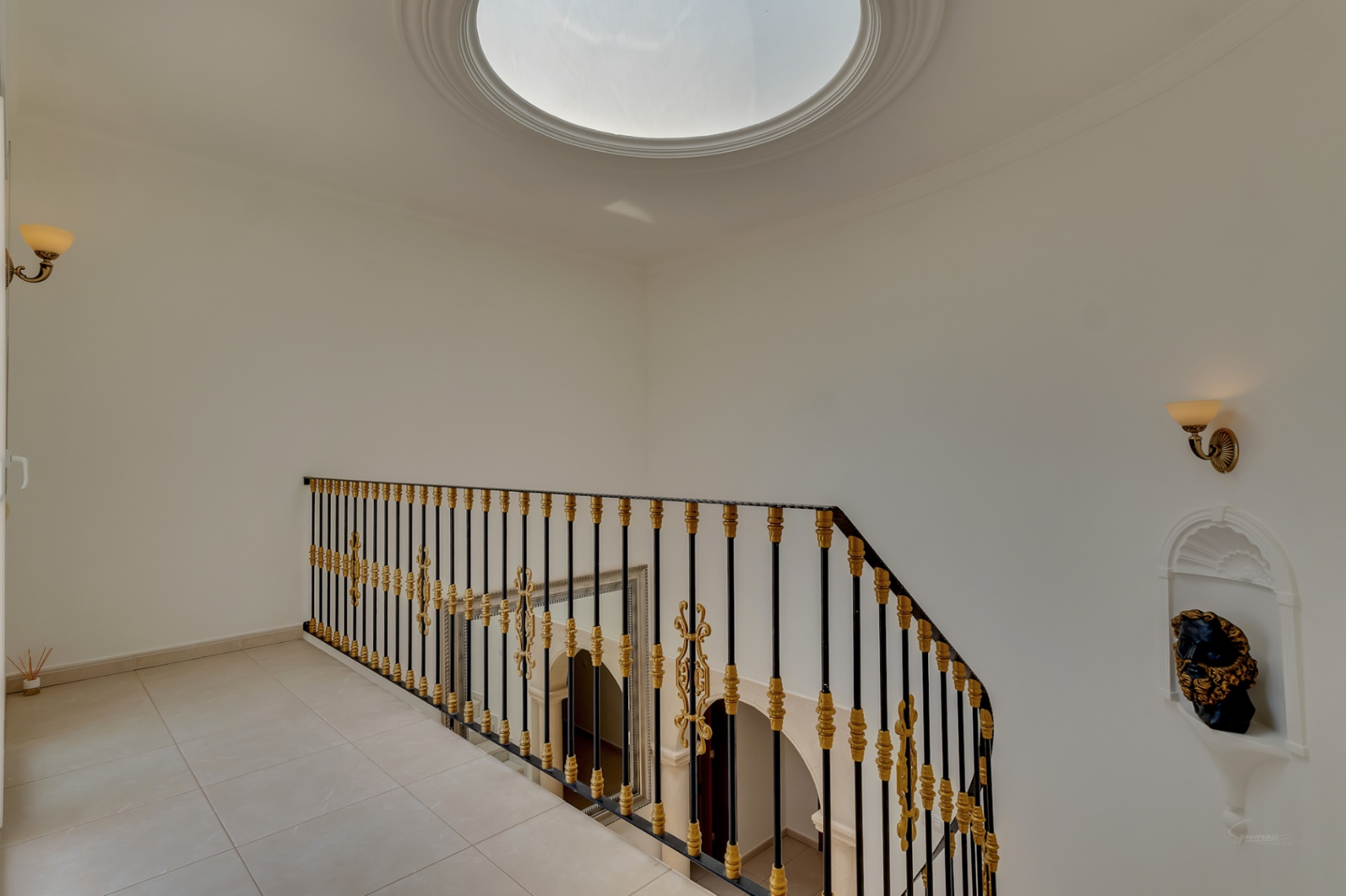 Palier supérieur avec coupole vitrée circulaire, balustrade en fer forgé aux détails dorés et niche murale décorative avec éclairage d'applique.
