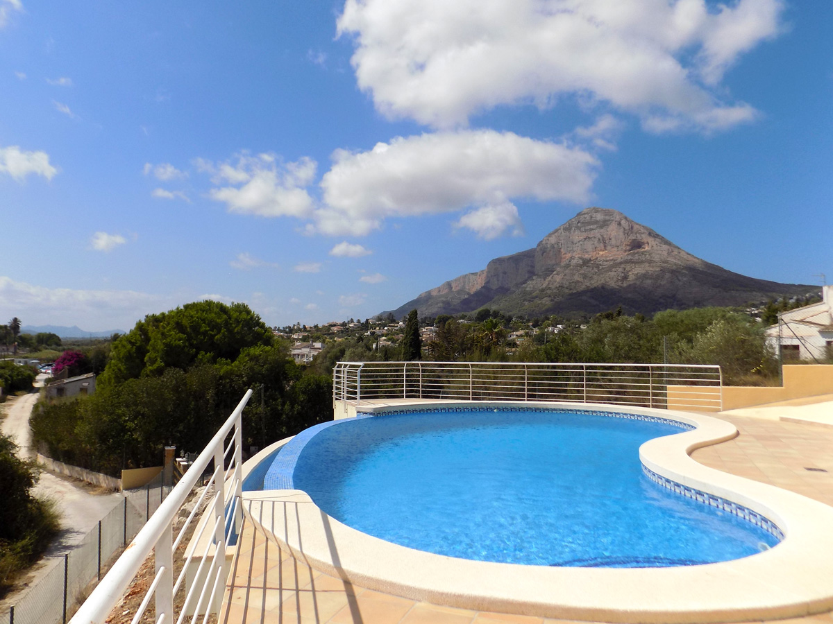 Piscine à débordement aux formes courbes et terrasse carrelée, offrant une vue panoramique sur le mont Montgó et la végétation environnante.