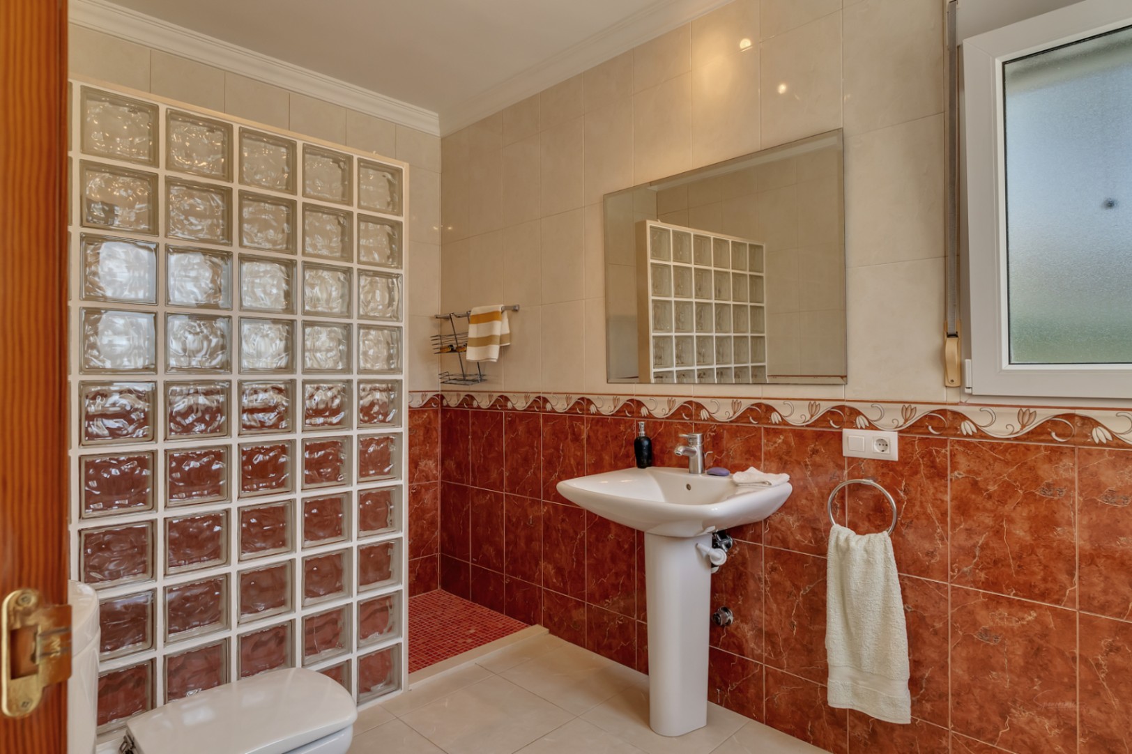 Salle de bain équipée d'un lavabo sur colonne, d'un grand miroir et d'une douche à l'italienne avec paroi en briques de verre et carrelage rouge marbré.