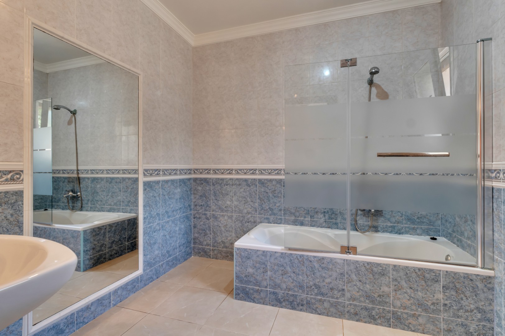 Salle de bains avec baignoire, paroi de douche en verre, carrelage mural bleu et blanc avec frise décorative et un grand miroir mural.
