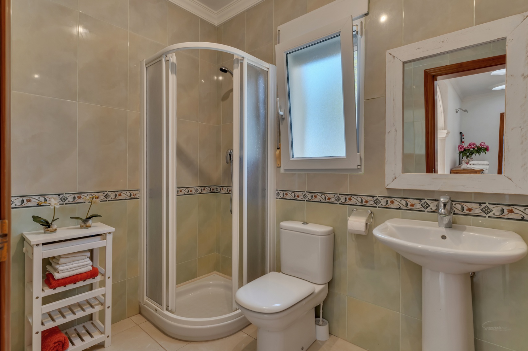 Salle d'eau équipée d'une cabine de douche d'angle, d'un lavabo sur colonne et de murs carrelés avec frise décorative et fenêtre apportant de la lumière.
