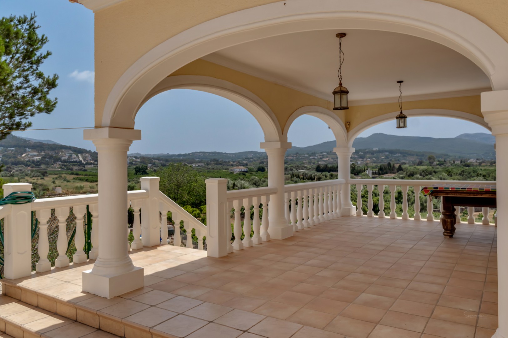 Terrasse couverte avec arches, colonnes blanches et balustrade. Sol en grès, luminaires suspendus et vue panoramique sur la vallée et les montagnes.
