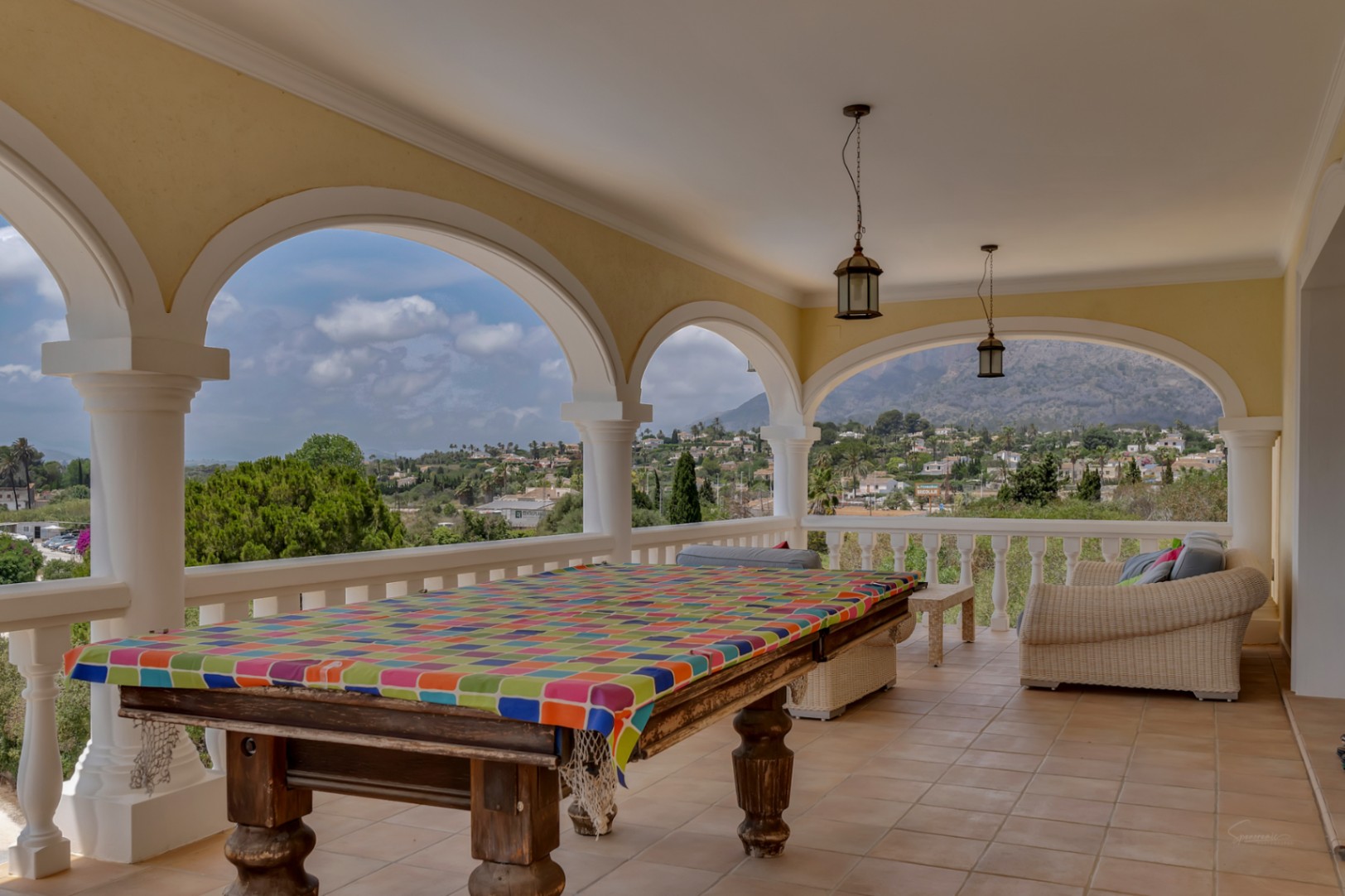 Terrasse couverte avec arches, table de billard et coin salon, offrant une vue panoramique sur la vallée de Jávea et le Montgó.