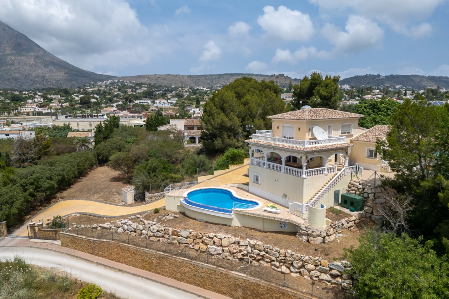 Vue aérienne de la villa avec piscine incurvée, terrasses à arcades et balustrades blanches, offrant une vue panoramique sur le Montgó et la vallée.