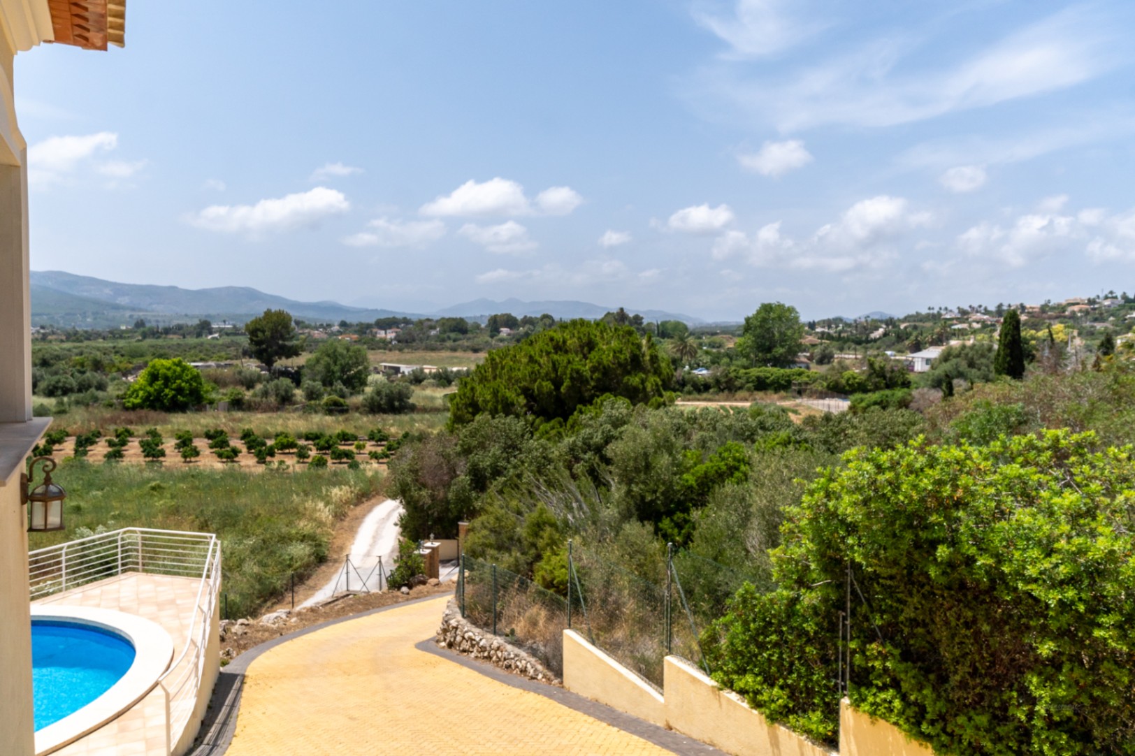 Vues panoramiques sur la vallée de Jávea et les orangeraies, illustrant l'allée pavée et la terrasse de la piscine circulaire avec ses garde-corps blancs.