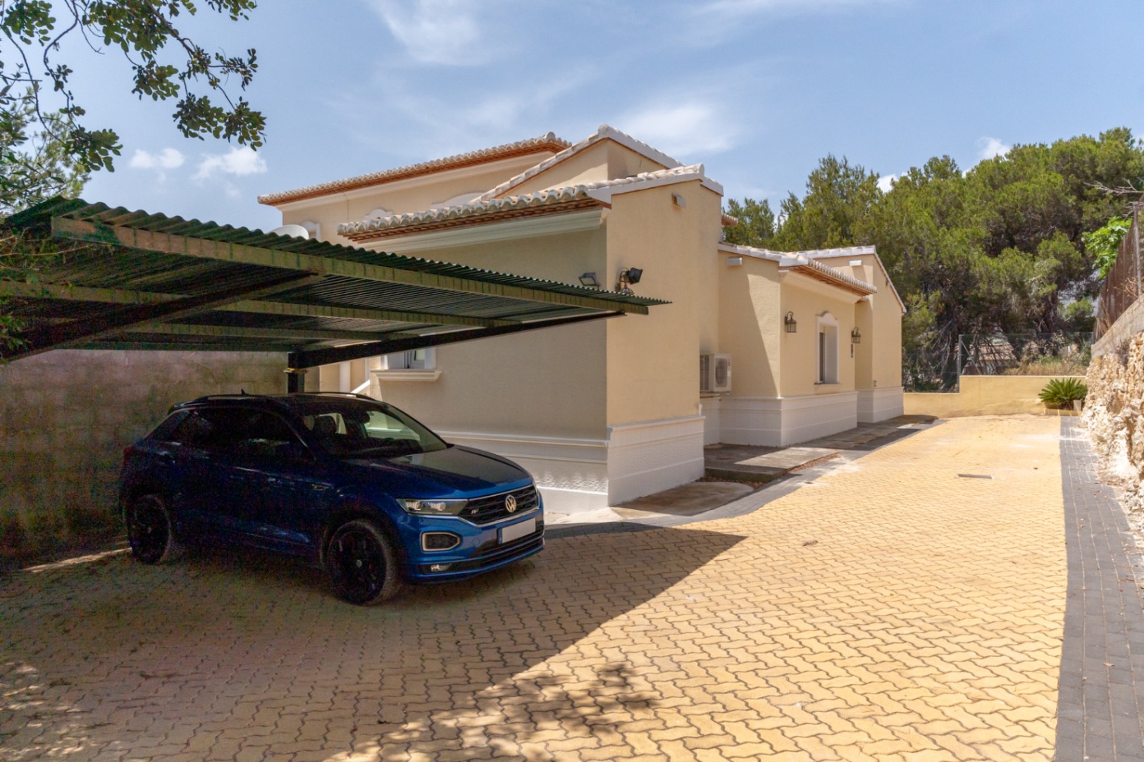 Zone de stationnement extérieure avec pavés autobloquants jaunes et un abri de voiture couvert situé à côté de la façade latérale de la villa.