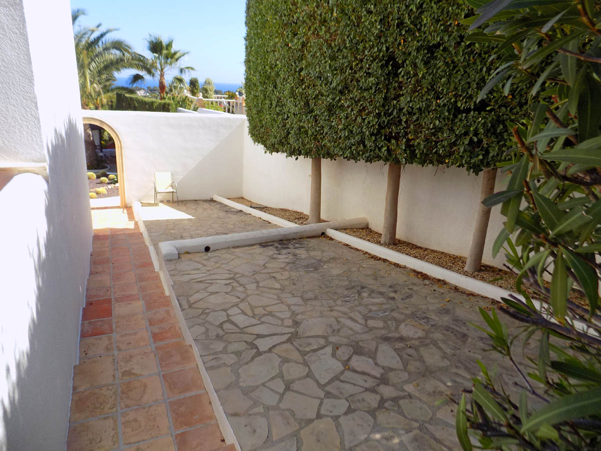 Allée extérieure avec pavage en pierre naturelle, murs blancs et arbres taillés avec précision, offrant une vue sur la mer à travers une arche.