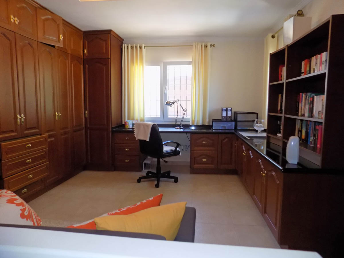 Bureau fonctionnel avec de grands placards intégrés en bois, un plan de travail en L de couleur sombre et une bibliothèque sous lumière naturelle.