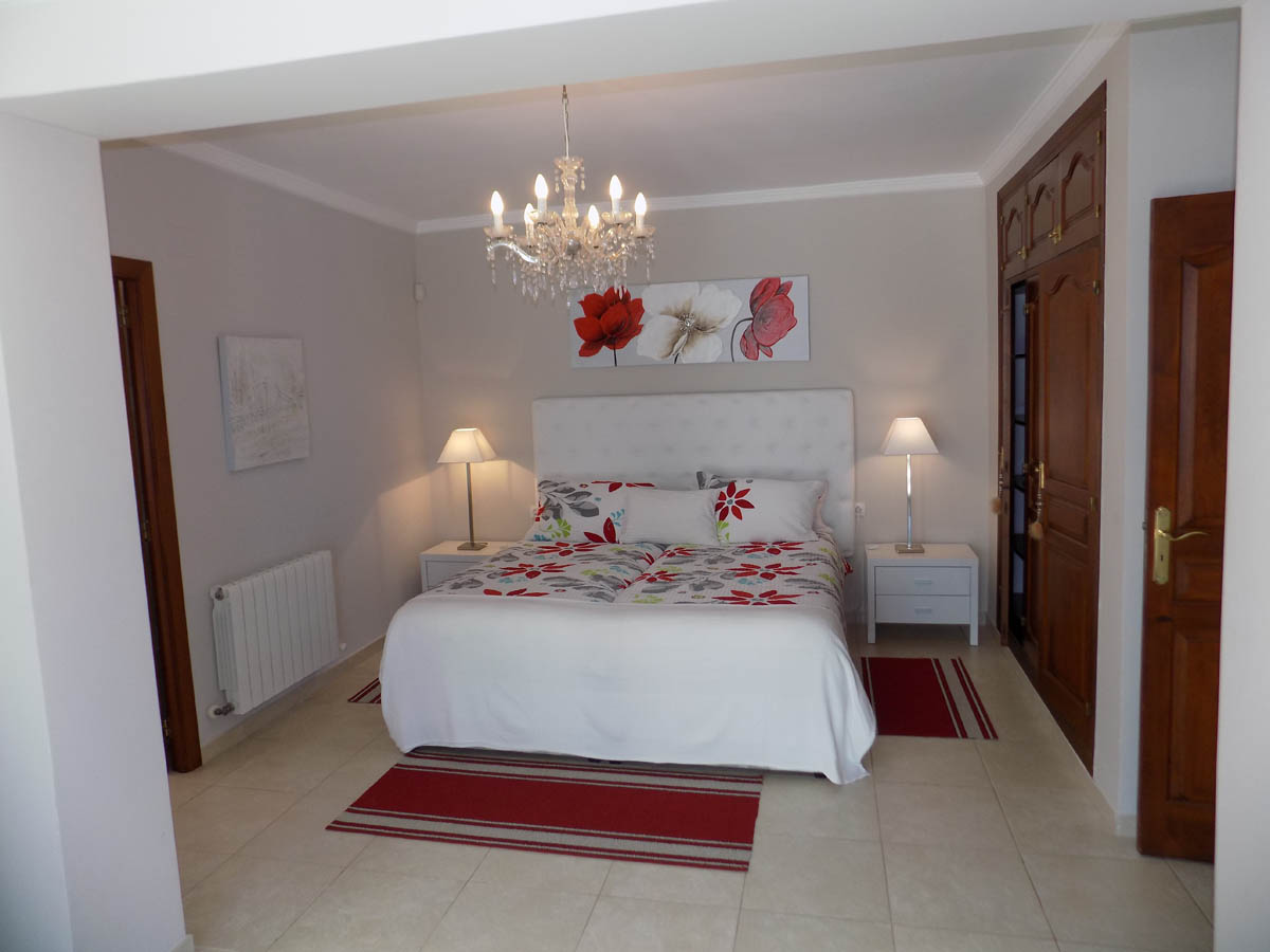 Chambre avec placards en bois intégrés, tête de lit capitonnée blanche, lustre en cristal et sol en carrelage clair avec radiateur.