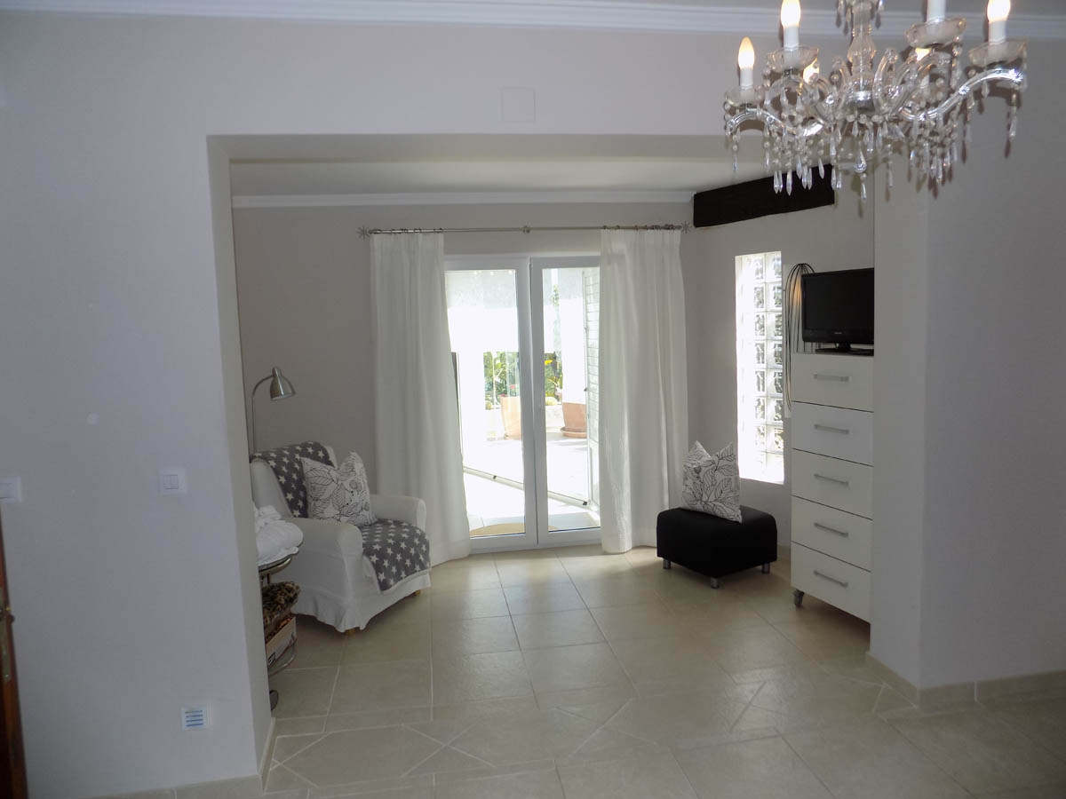 Chambre principale avec coin salon, carrelage géométrique clair et accès terrasse. Équipée d'une commode blanche et d'une fenêtre en briques de verre.