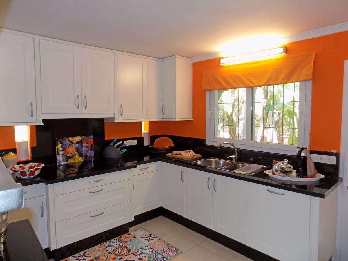 Cuisine équipée avec placards blancs, plans de travail en granit noir et murs orange vif. Comprend un double évier en inox et une fenêtre offrant une vue sur le jardin.