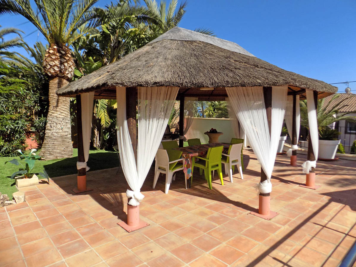 Espace de détente extérieur avec gazebo en chaume, rideaux blancs élégants et coin repas sur terrasse en terre cuite entourée de palmiers.