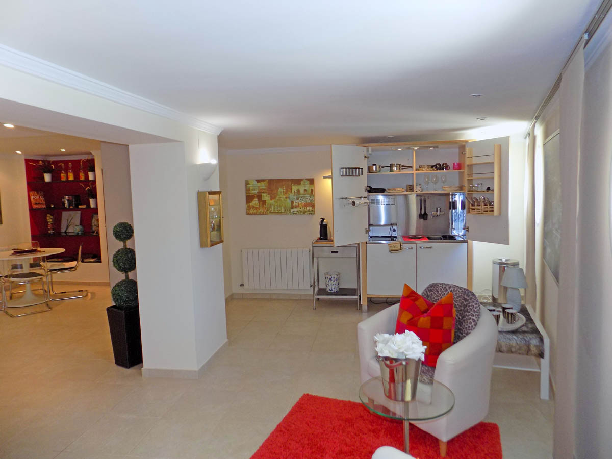 Espace de vie au rez-de-chaussée avec kitchenette compacte intégrée, coin salon avec fauteuil blanc et tapis rouge, et salle à manger avec étagères murales.