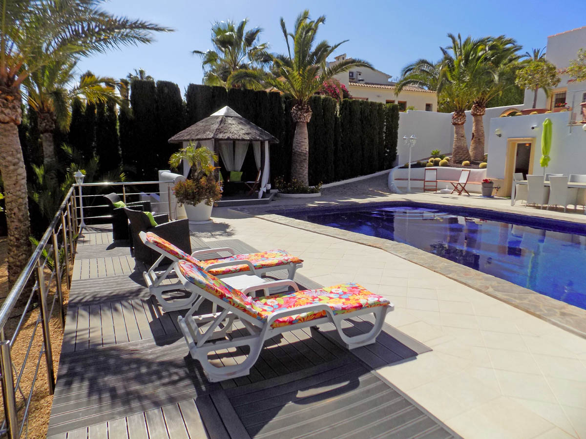 Espace piscine avec terrasse en pierre et bois composite, chaises longues, gazebo en chaume et jardin méditerranéen avec palmiers et haies denses.