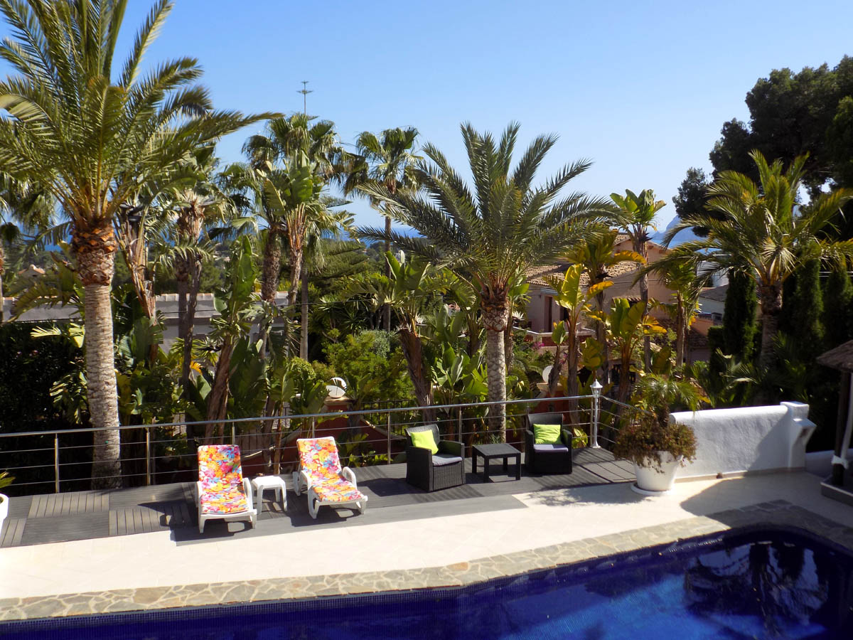 Terrasse surélevée avec piscine, solarium en bois composite et vue sur mer à travers une végétation méditerranéenne luxuriante et des palmiers.