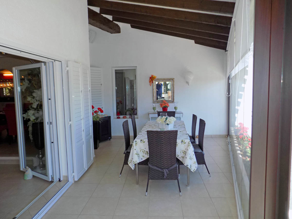 Terrasse vitrée avec poutres en bois apparentes, coin repas pour six personnes et accès direct à l'intérieur par des portes coulissantes blanches.
