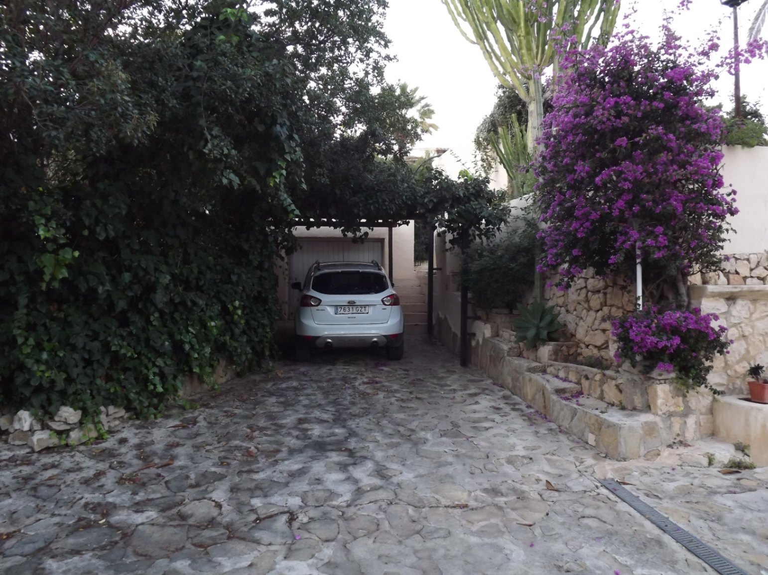 Allée en pierre naturelle menant au garage, bordée de végétation méditerranéenne luxuriante, de bougainvilliers et de murs en pierre sèche.