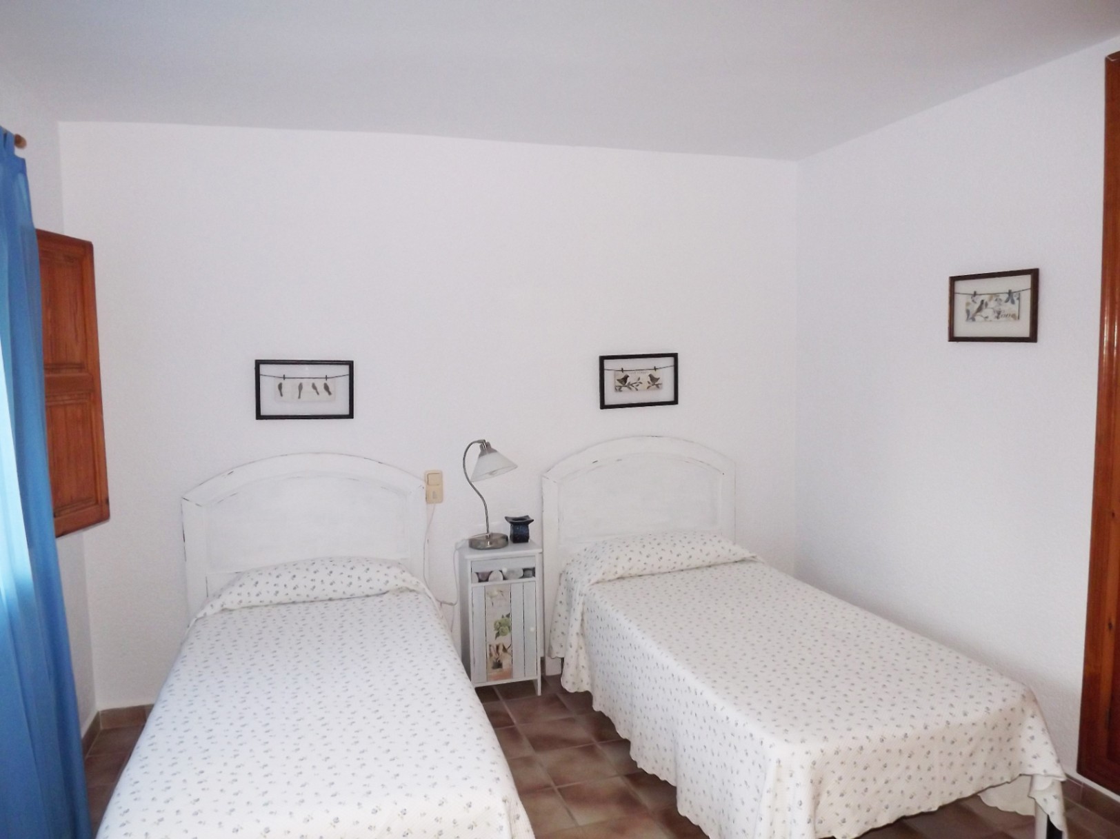 Chambre avec deux lits simples, têtes de lit blanches cintrées et sol en terre cuite. Elle dispose de volets en bois et d'un éclairage naturel.