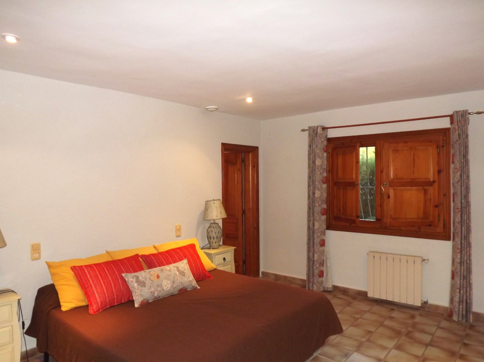 Chambre double avec sol en grès, fenêtre en bois avec volets intérieurs et radiateur. Équipée d'un éclairage intégré au plafond et de menuiseries en bois massif.