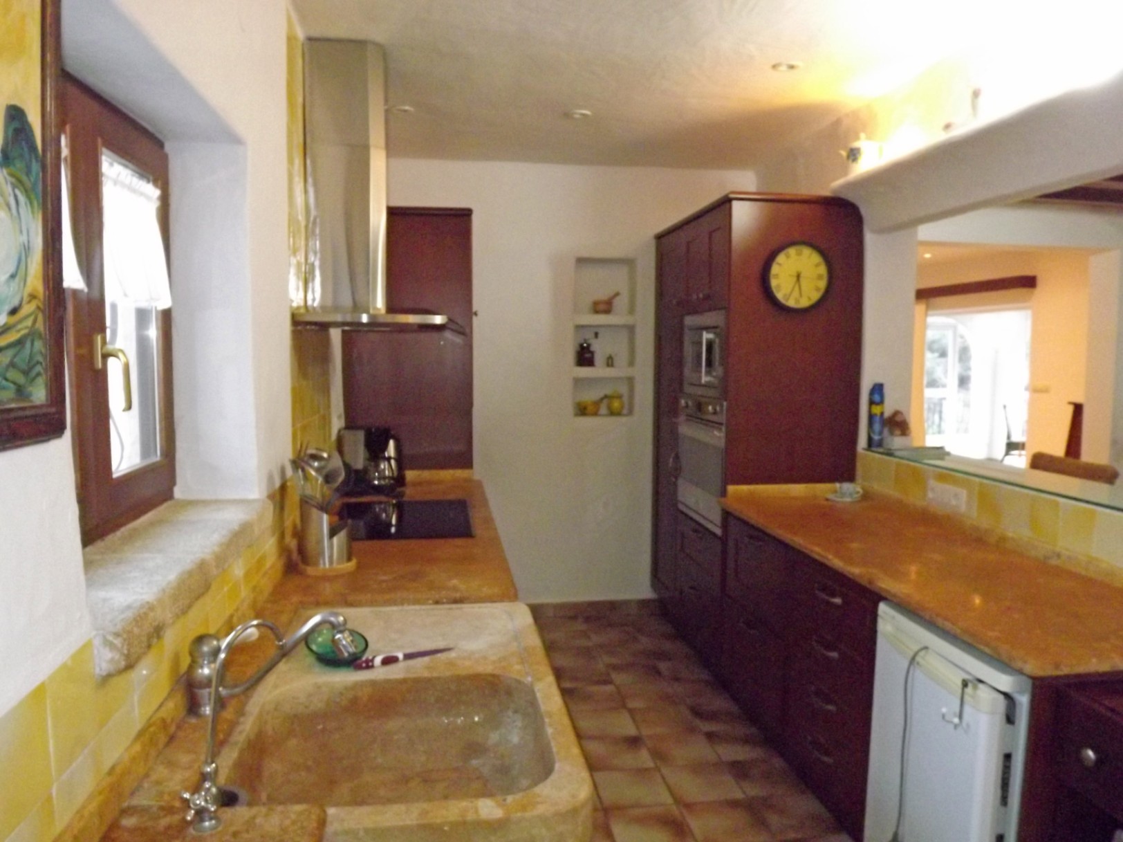 Cuisine rustique avec évier en pierre taillée, meubles en bois sombre et crédence jaune. Elle offre un plan de travail en pierre et une ouverture vers le séjour.