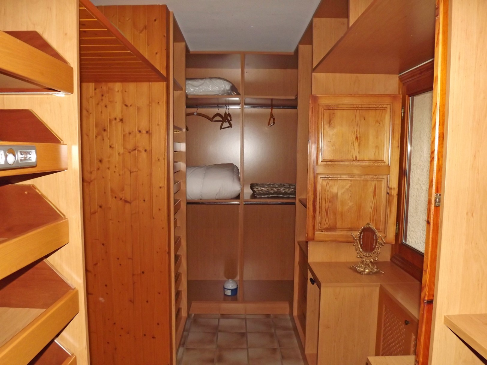 Dressing spacieux avec étagères en bois sur mesure, tringles de suspension et sol en grès. Une fenêtre avec volet intérieur en bois assure la ventilation de l'espace.