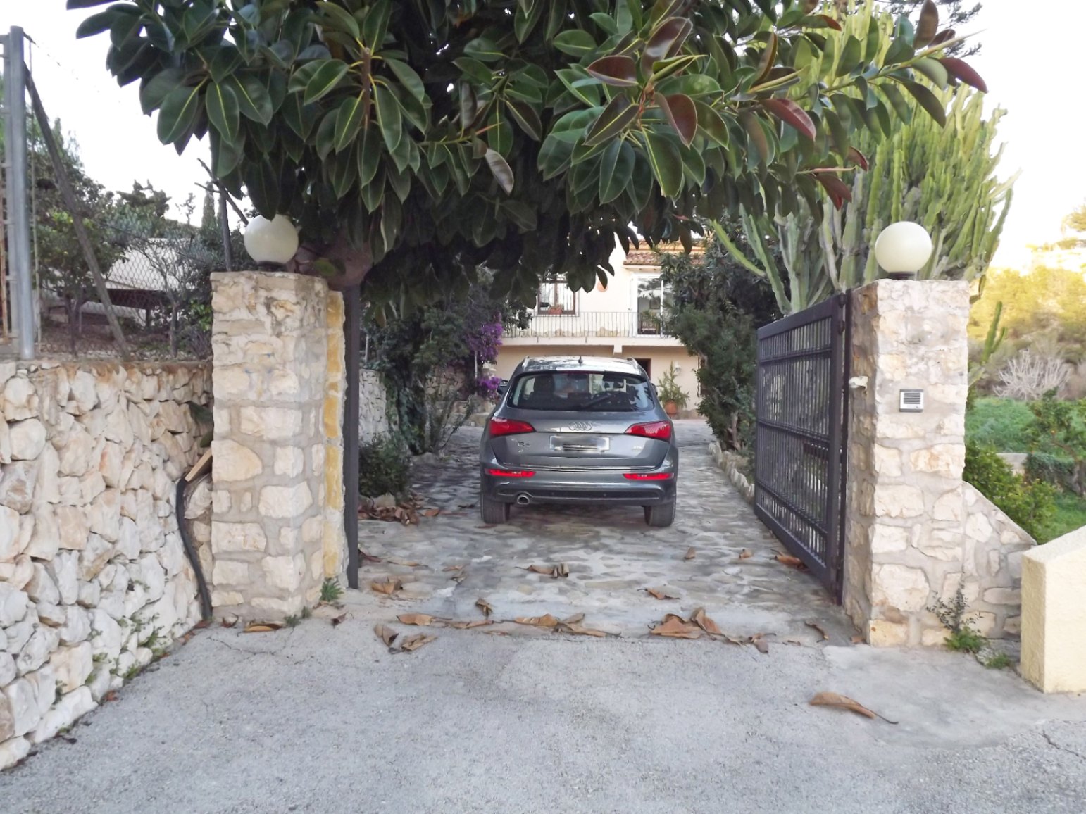 Entrée principale avec murs en pierre naturelle, portail métallique et allée pavée entourée d'une végétation méditerranéenne luxuriante.