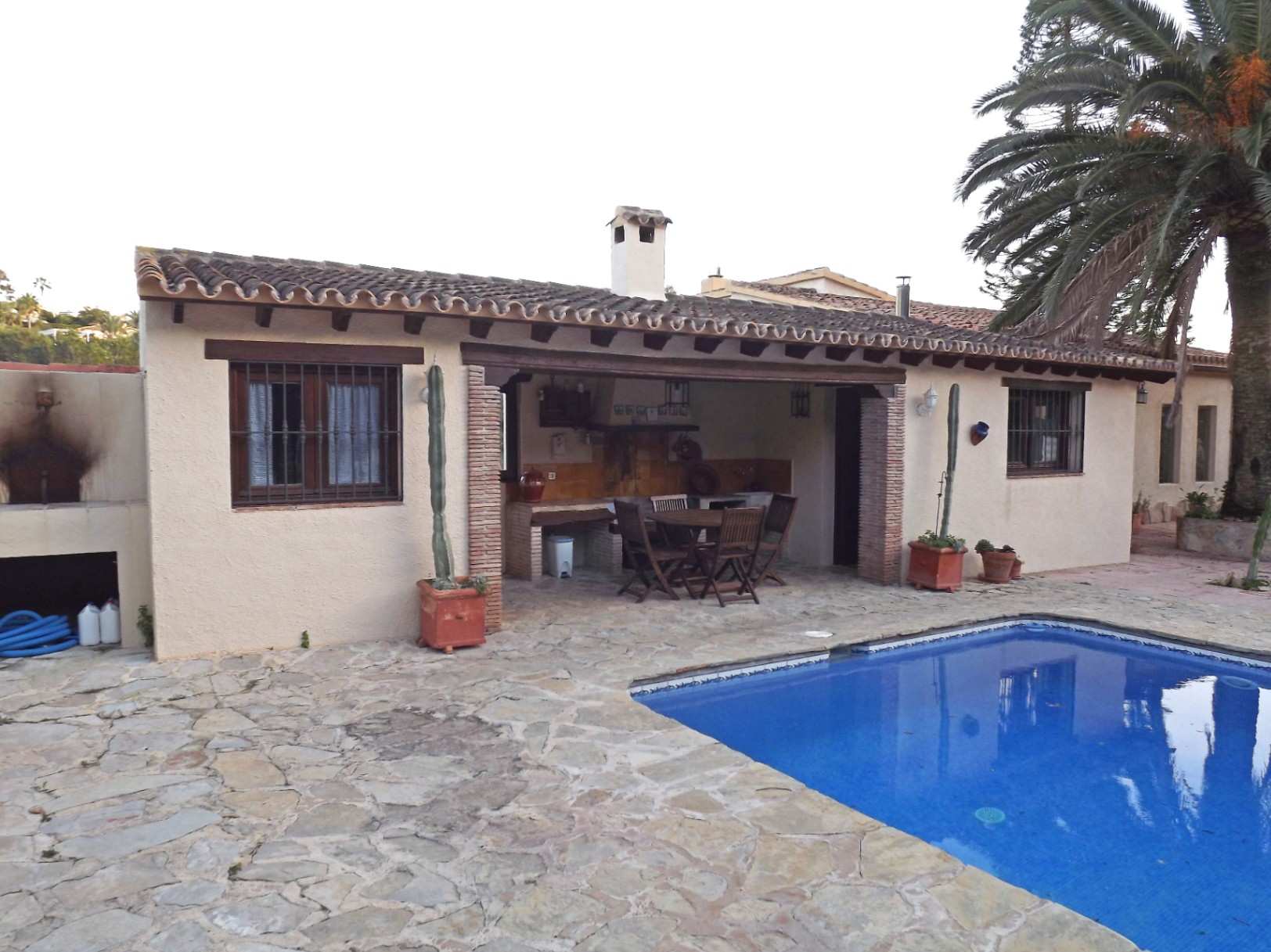 Espace extérieur avec piscine carrelée, vaste terrasse en pierre naturelle et porche couvert avec poutres en bois et barbecue intégré.