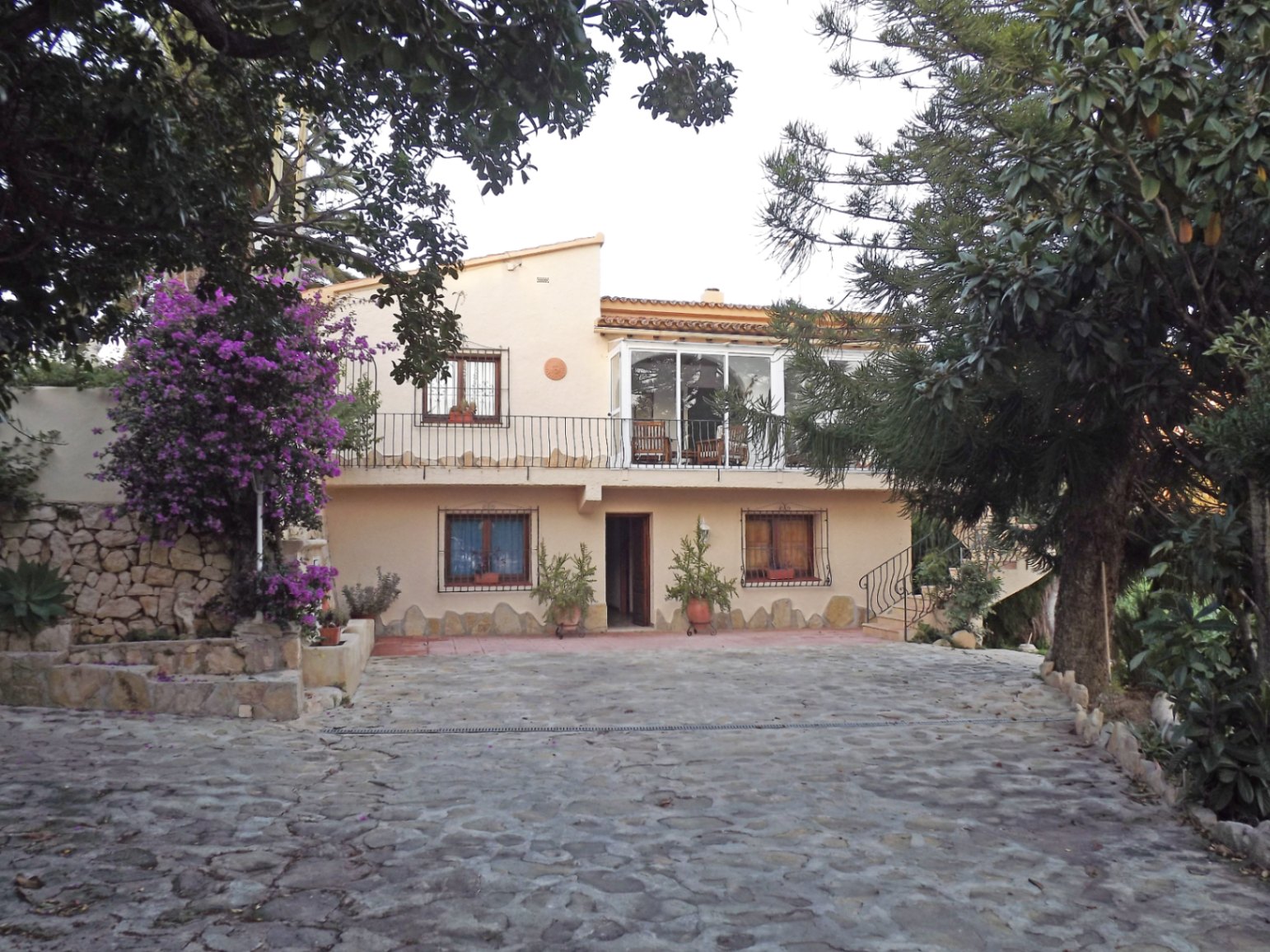 Façade principale de la villa avec une entrée pavée de pierres, une terrasse vitrée à l'étage et des détails en fer forgé entourés de bougainvilliers.
