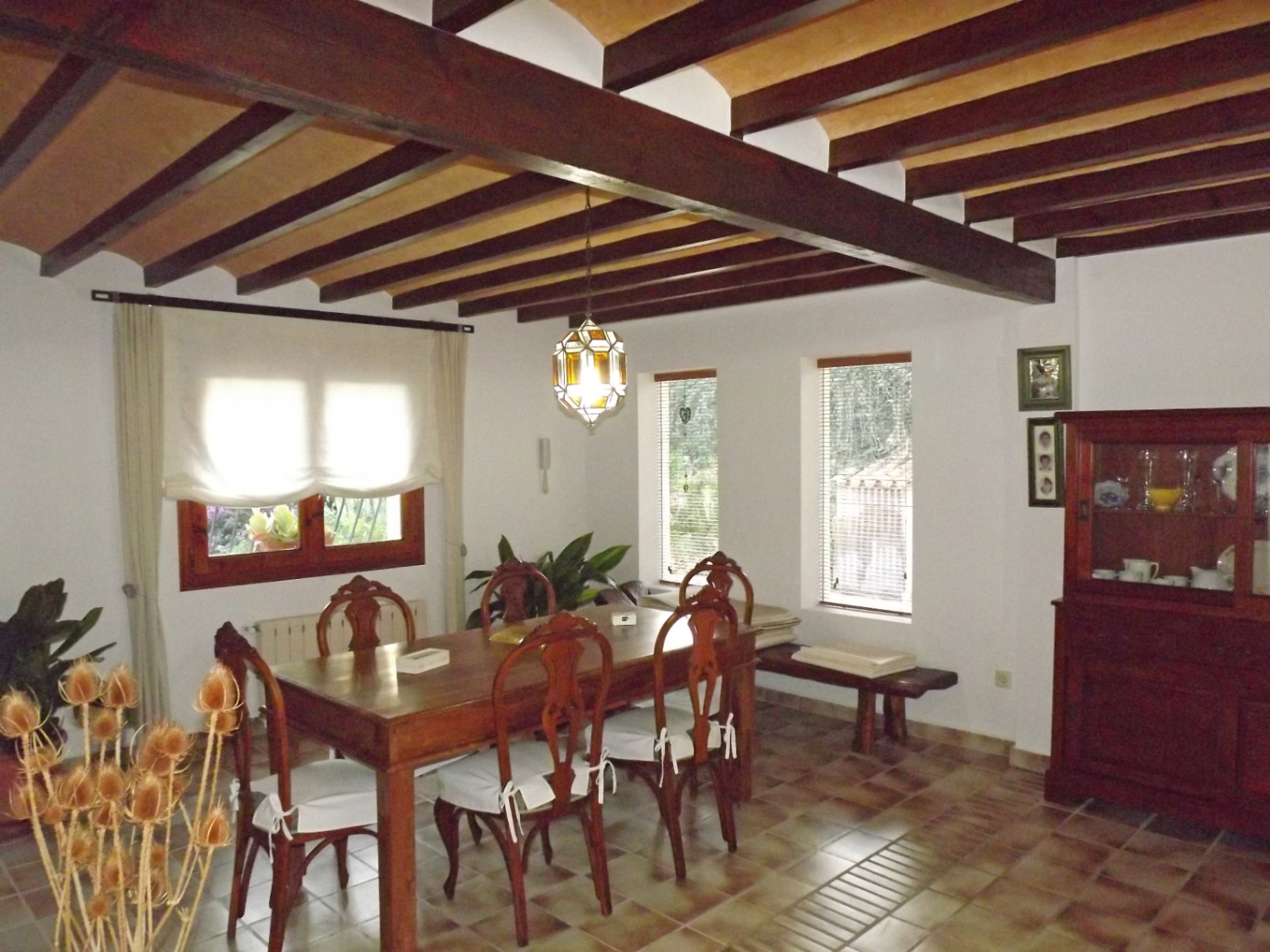 Salle à manger rustique avec poutres apparentes en bois sombre, table massive et sol carrelé. La pièce dispose d'un buffet classique et d'un éclairage naturel constant.