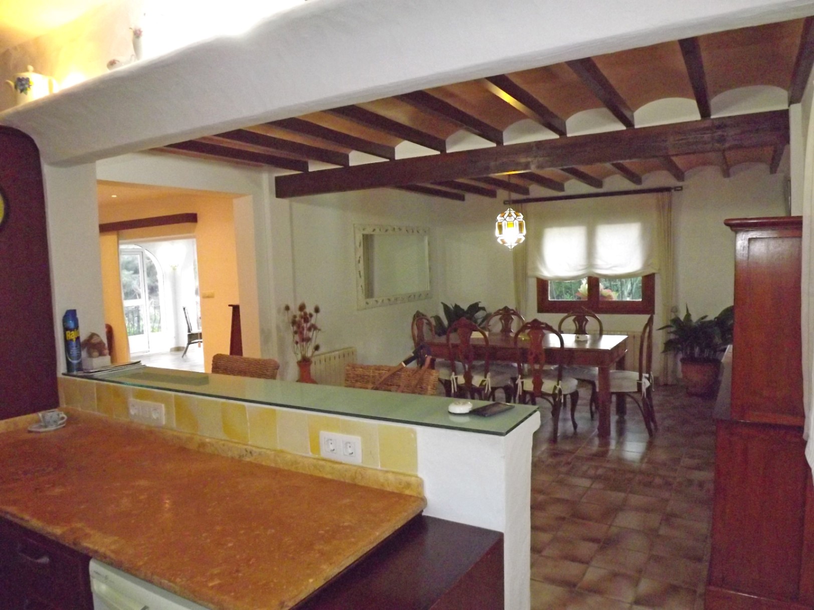 Salle à manger rustique avec poutres apparentes et voûtes en terre cuite, ouverte sur la cuisine par un comptoir en pierre et une étagère en verre.