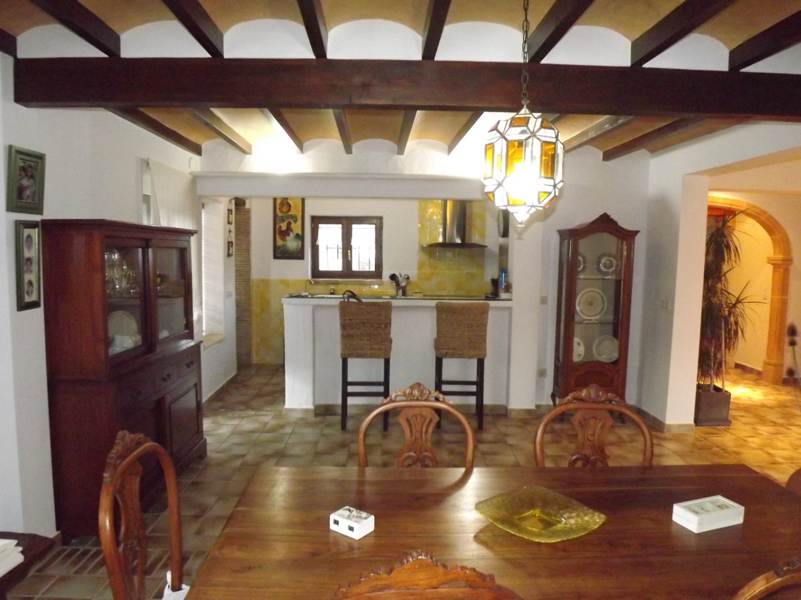 Salle à manger rustique avec poutres apparentes, table en bois massif et cuisine ouverte équipée d'un bar avec tabourets en osier.