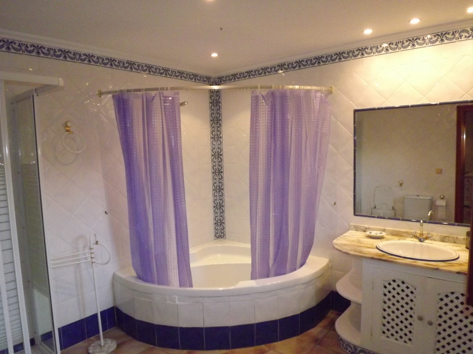 Salle de bain avec baignoire d'angle, rideaux violets, meuble-vasque en marbre avec portes en treillis et carrelage blanc aux bordures bleues.