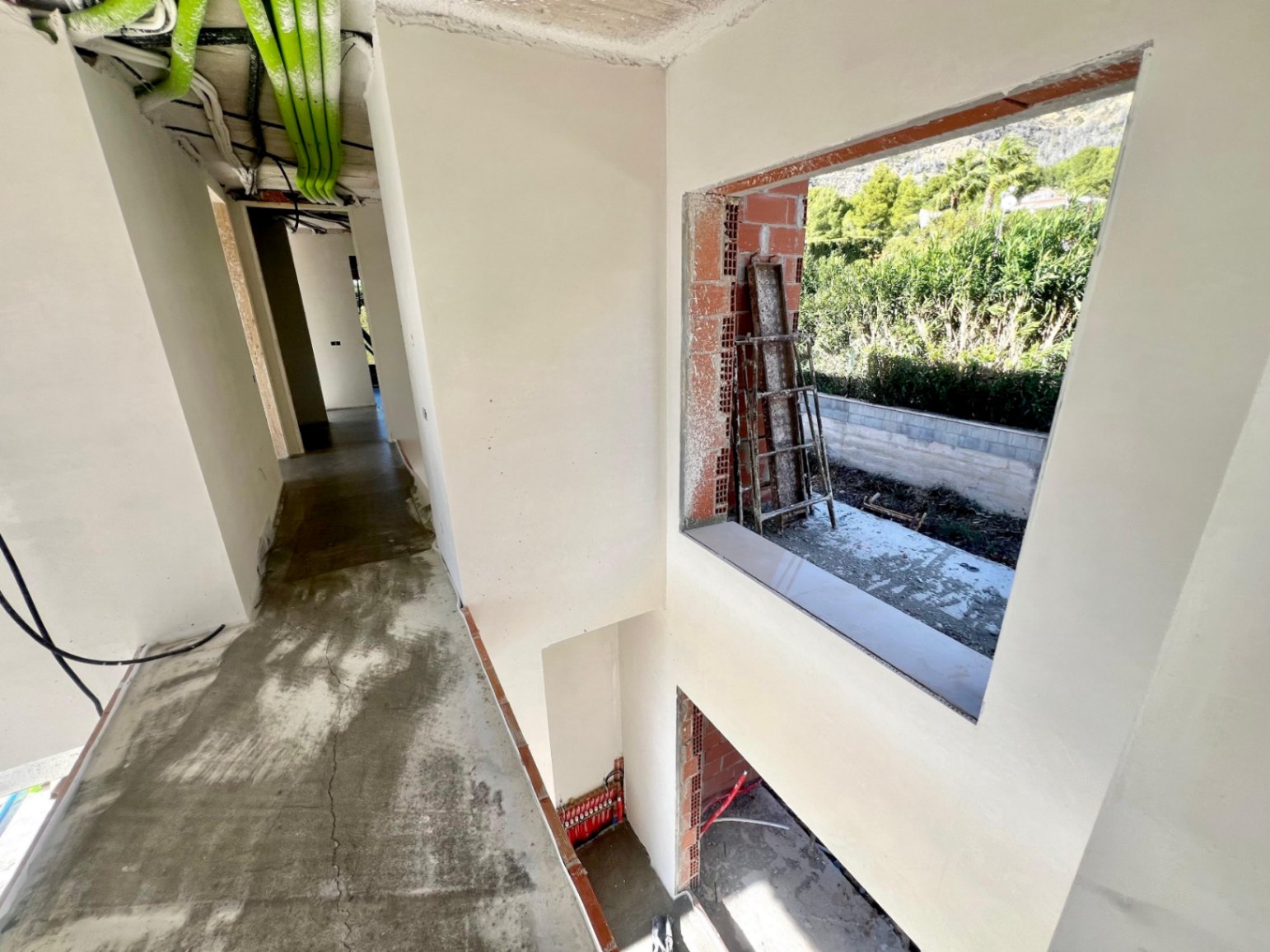 Couloir intérieur en phase de gros œuvre, présentant les gaines techniques au plafond et une ouverture offrant une vue sur la végétation environnante.