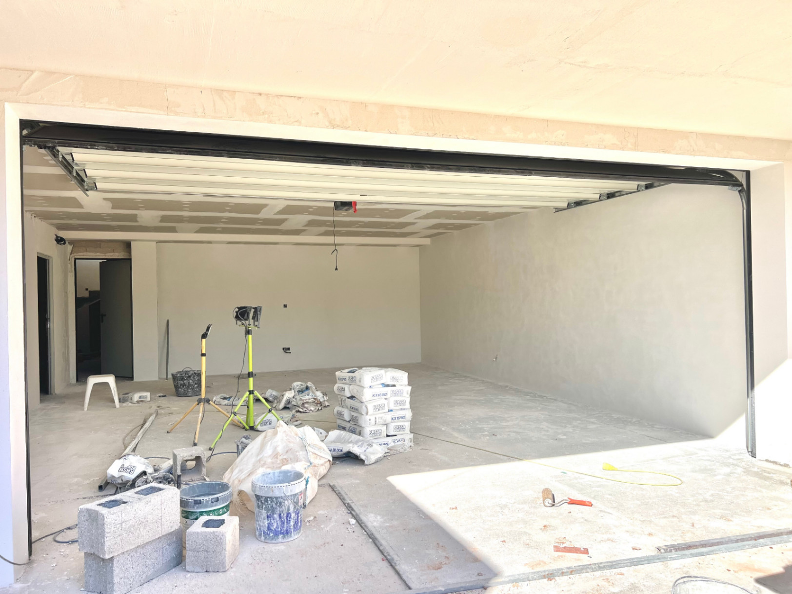 Grand garage en cours de construction avec porte sectionnelle blanche, murs enduits et plafonds en plaques de plâtre, idéal pour plusieurs véhicules.