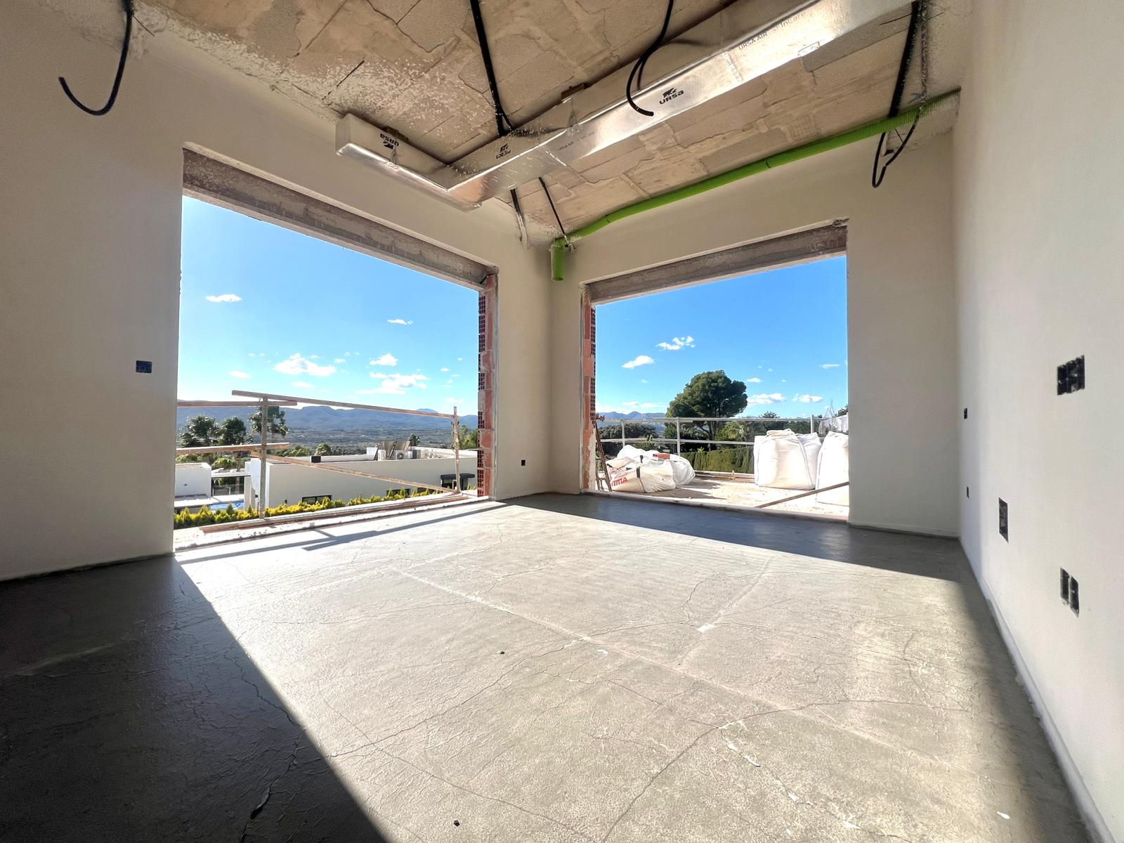 Intérieur en cours de construction avec de grandes baies vitrées, plafonds en béton, gaines de climatisation et vues dégagées sur les montagnes.