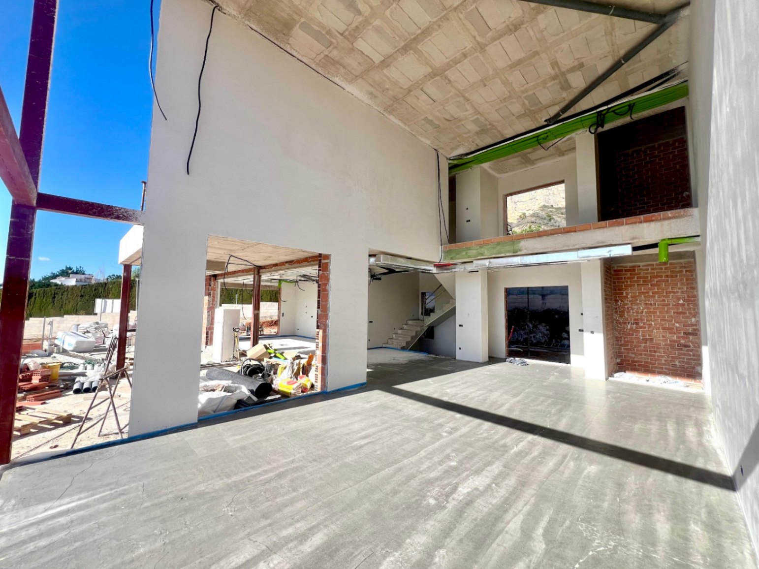 Vue intérieure de l'espace de vie à double hauteur en construction, avec mezzanine ouverte, escalier en béton et larges ouvertures pour baies vitrées.
