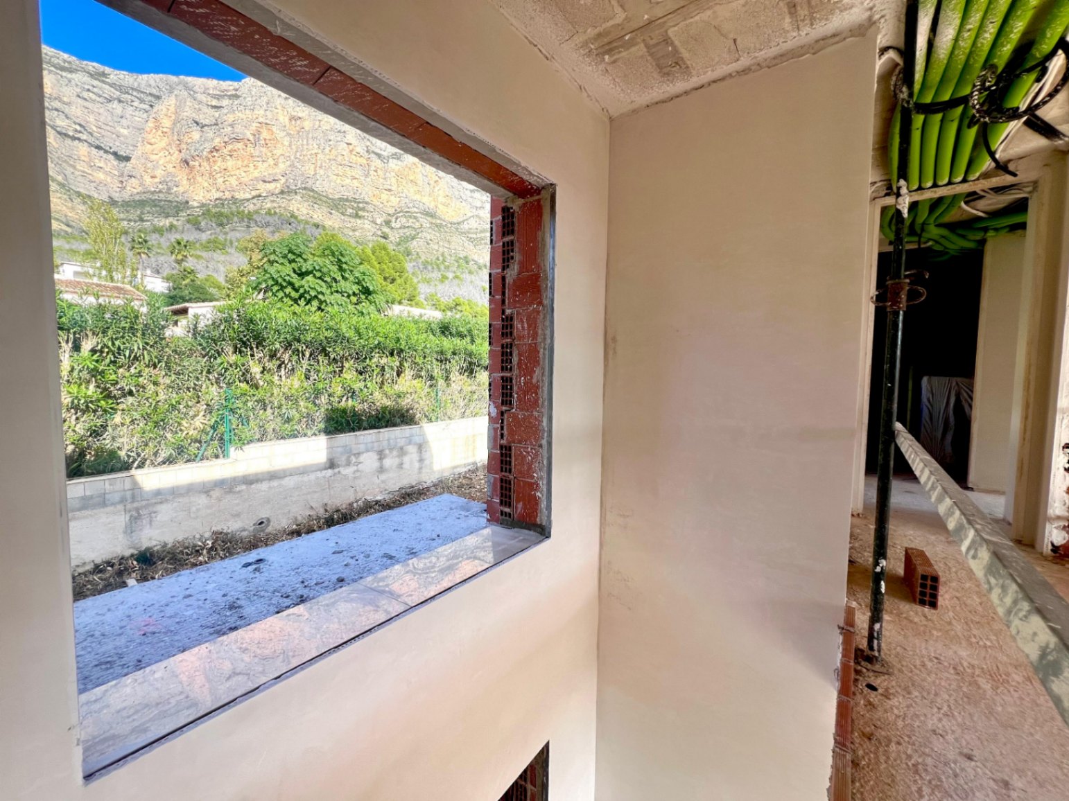 Vue intérieure en chantier avec une large ouverture sur le mont Montgó, révélant les gaines techniques au plafond et les murs en cours de finition.