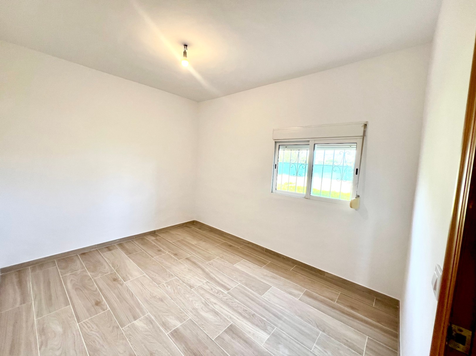 Chambre lumineuse avec sol en grès cérame imitation bois, murs blancs et fenêtre sécurisée par une grille ornementale et un volet roulant.