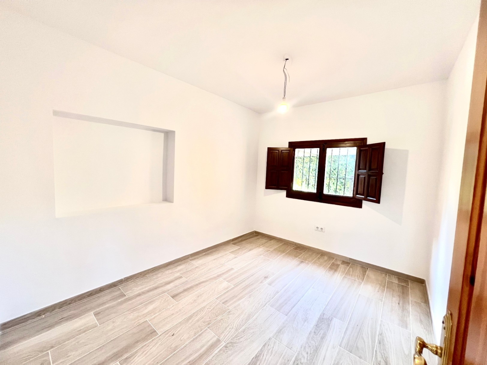 Chambre lumineuse avec sol en grès cérame imitation bois, niche murale intégrée et fenêtre équipée de volets intérieurs en bois sombre.