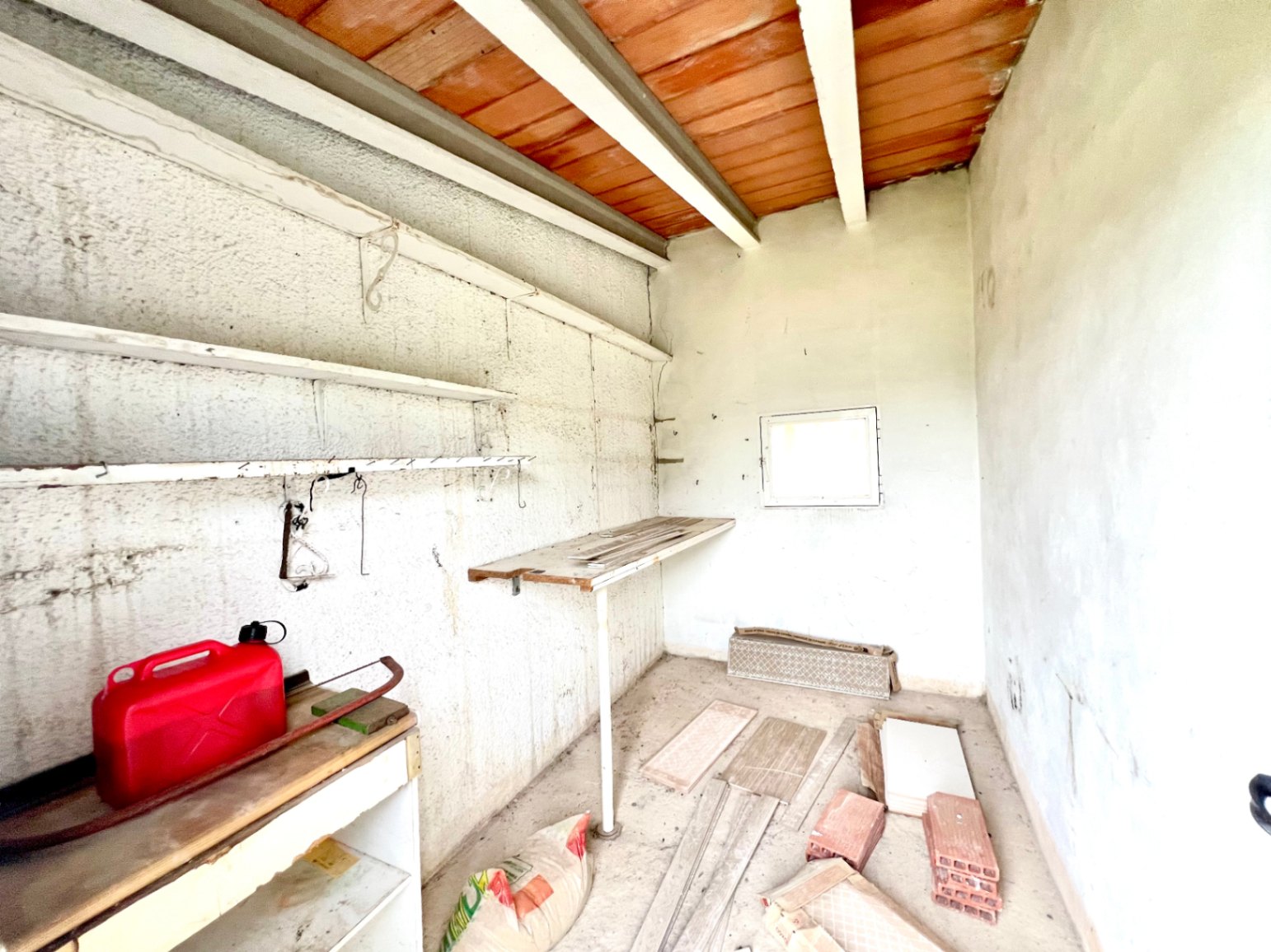 Espace de rangement ou atelier avec plafond à poutres apparentes, étagères murales et lumière naturelle apportée par une petite fenêtre carrée.