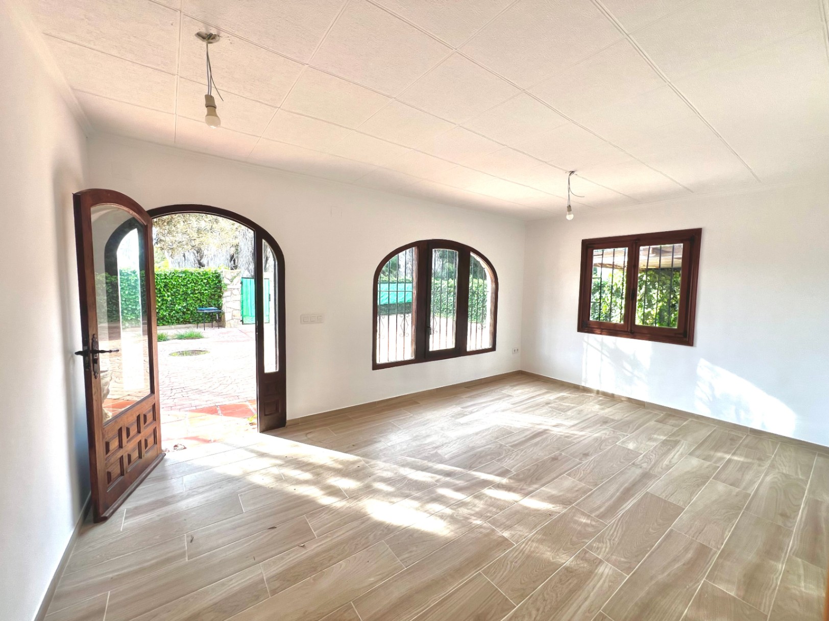 Espace de vie lumineux avec sol en grès imitation bois, fenêtres cintrées en bois avec grilles de sécurité et accès direct vers la terrasse extérieure pavée.