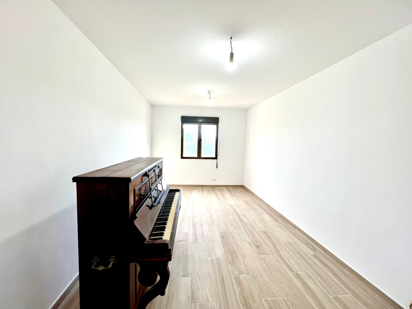 Pièce lumineuse avec murs blancs, sol en grès cérame imitation bois, un piano droit classique et une fenêtre à cadre sombre au fond.