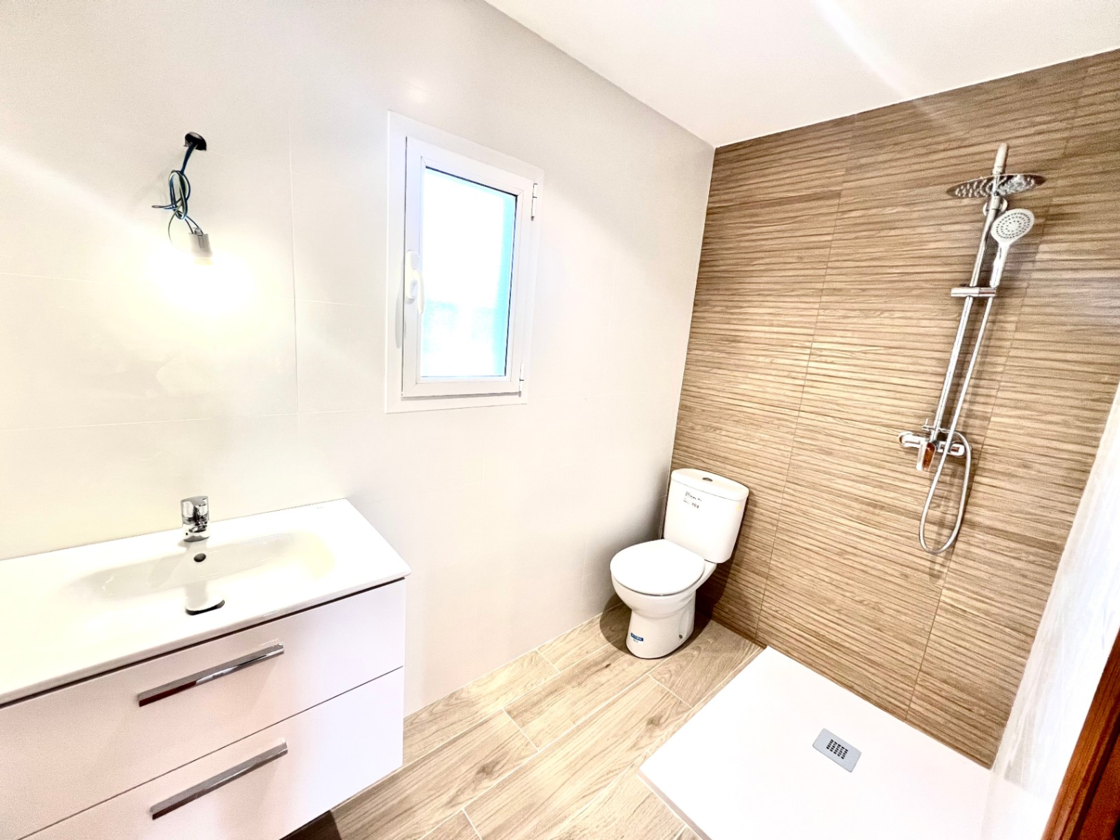 Salle de bain moderne avec douche à l'italienne, carrelage aspect bois et meuble vasque blanc. Une fenêtre en PVC apporte une luminosité naturelle à l'espace.