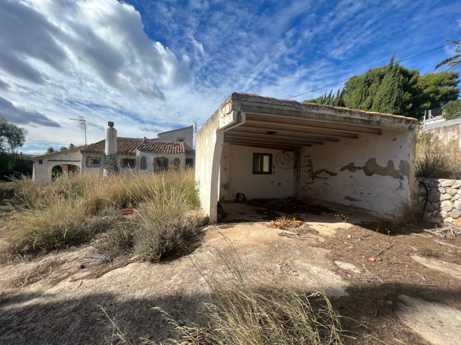 Abri de voiture indépendant et débarras à rénover, situés sur un vaste terrain avec végétation sauvage et vue sur la maison principale de style traditionnel.