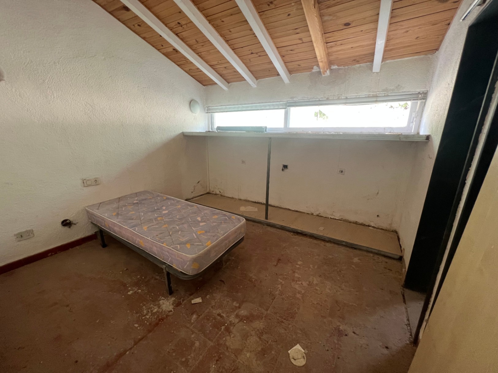 Chambre avec plafond incliné, poutres en bois apparentes et chevrons blancs. Elle dispose d'un sol en terre cuite d'origine et d'une fenêtre horizontale allongée.