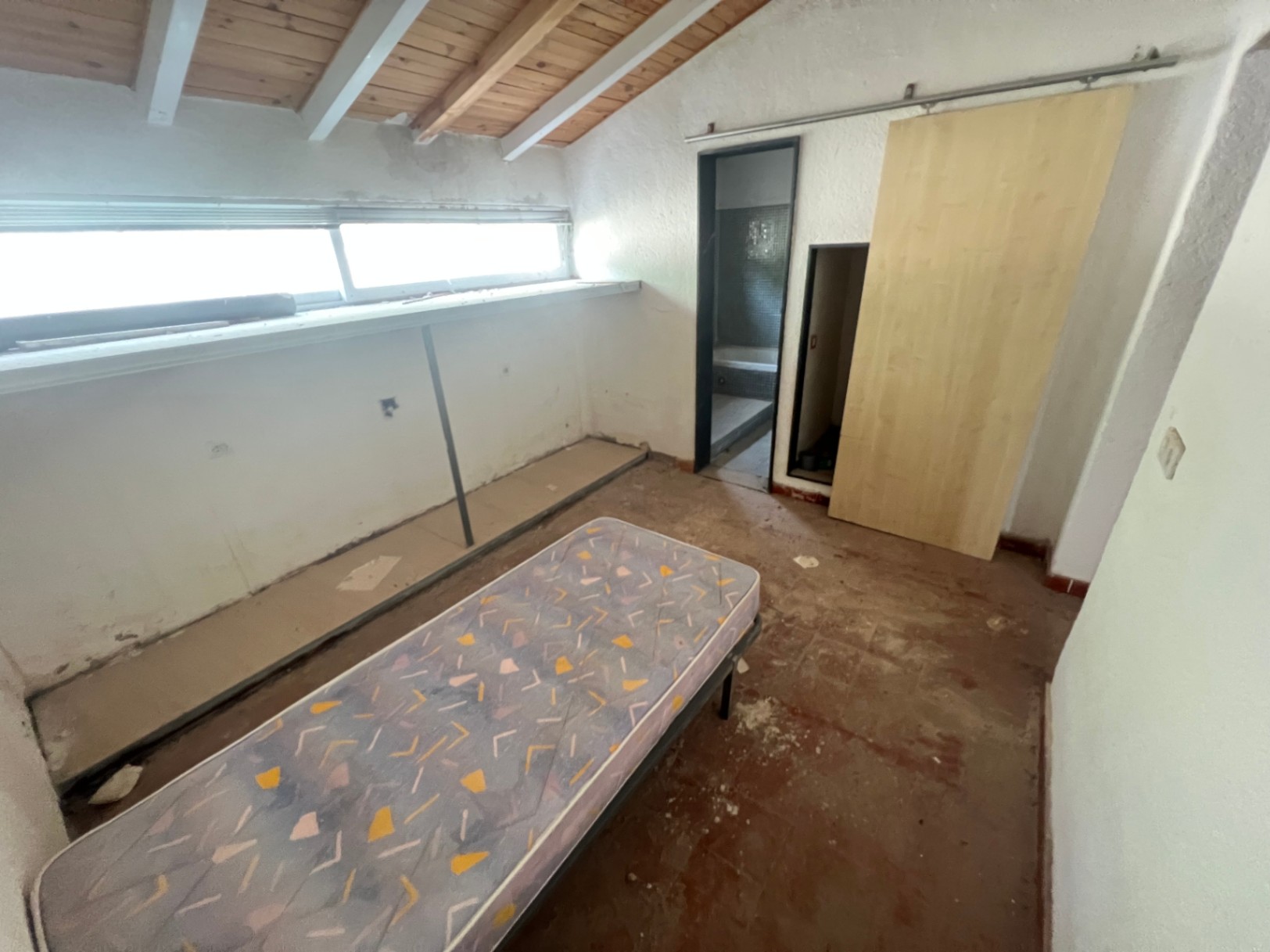 Chambre avec plafond mansardé en poutres de bois, fenêtre horizontale et porte coulissante ouvrant sur une salle de bain attenante à rénover entièrement.